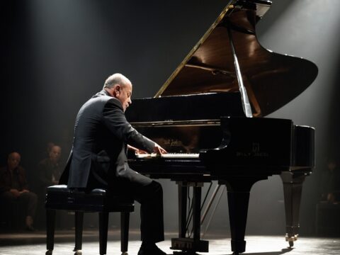Billy Joel sits at a grand piano on a dimly lit stage, spotlighted.
