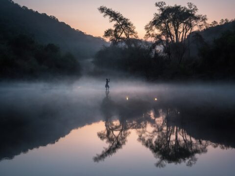 Faint, glowing shapes resembling dancers reflected on a mist-covered river at dusk.