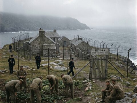 Early color photo of a guarded island colony with stone barracks, fences, and exiled men.